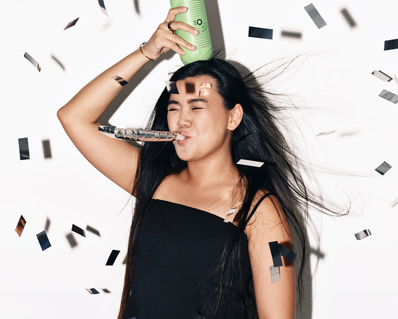 Donna in un vestito nero festeggia con una tromba da festa e una bomboletta spray verde, circondata da coriandoli.