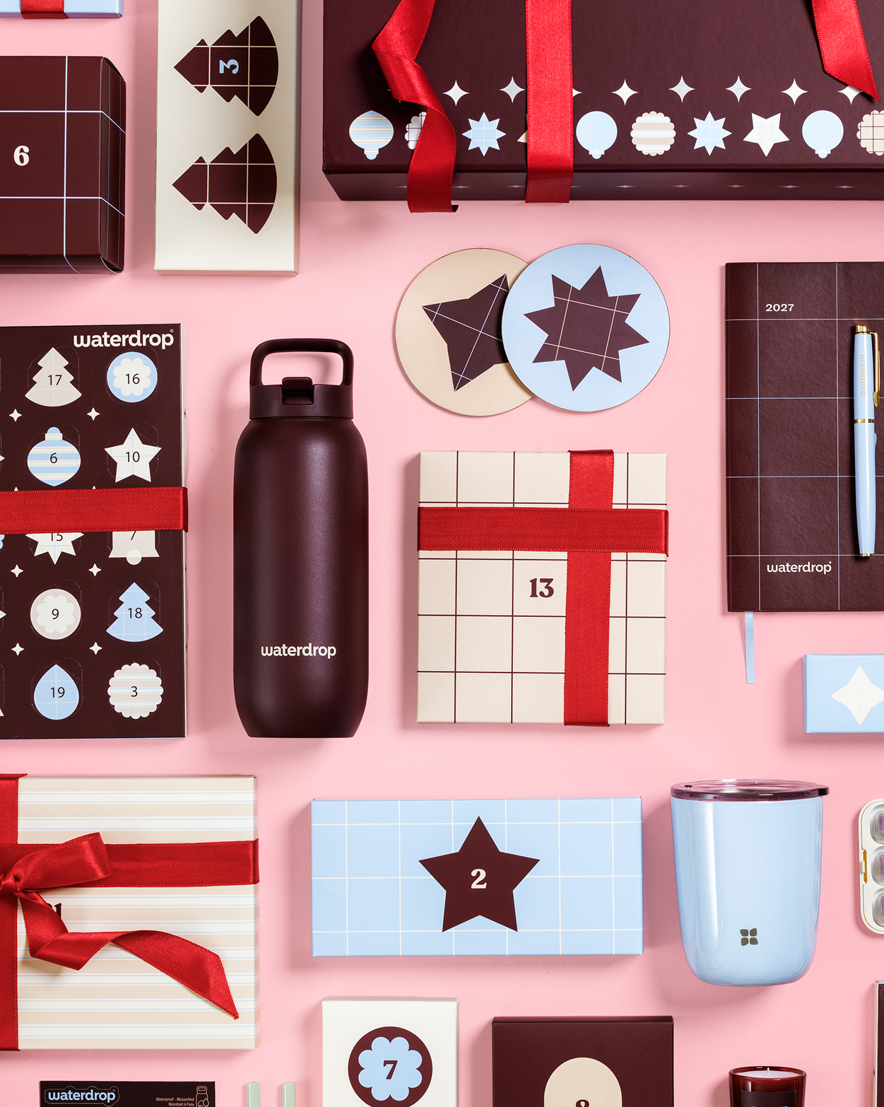 Scatole regalo assortite, calendario dell'avvento, una bottiglia d'acqua, una tazza e altri regali incartati su uno sfondo rosa.