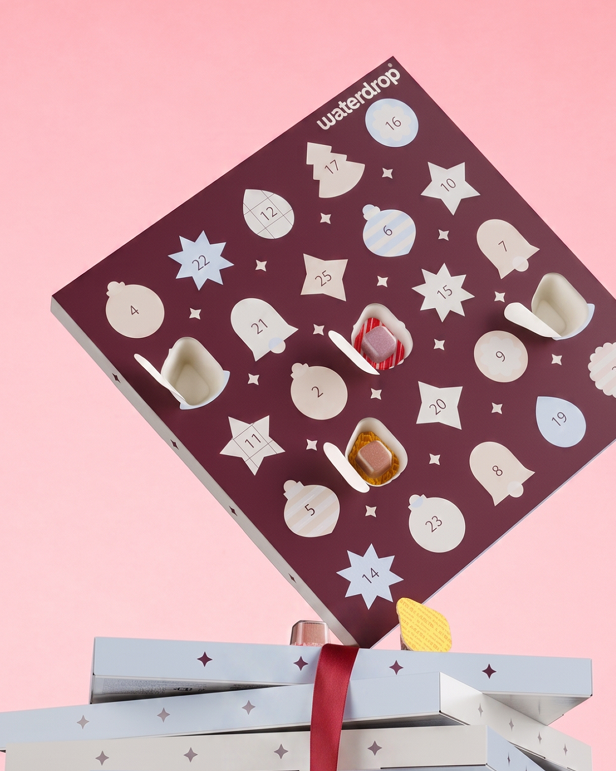 Calendario dell'Avvento viola con porte a forma di stelle numerate su sfondo rosa, posto sopra due scatole regalo incartate.