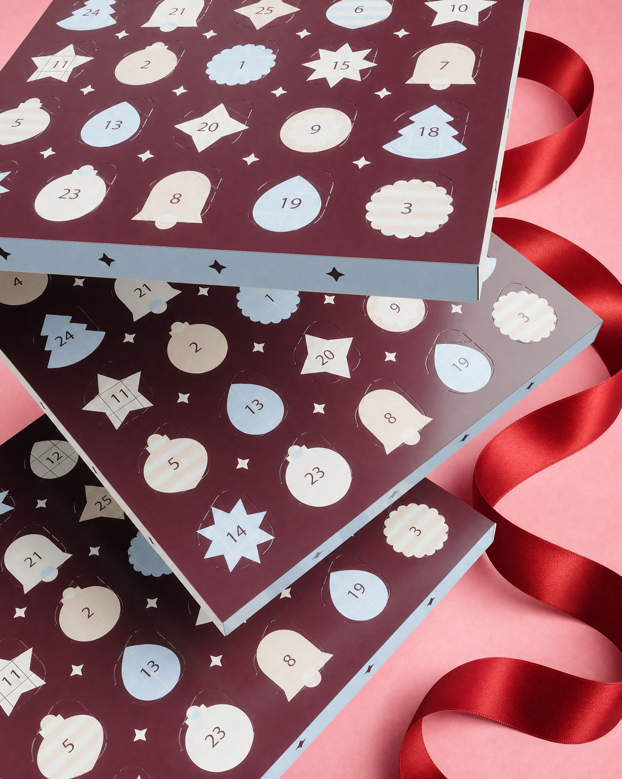 Tre scatole di calendario dell'Avvento impilate con sportelli numerati e decorazioni, annodate con un nastro rosso su sfondo rosa.