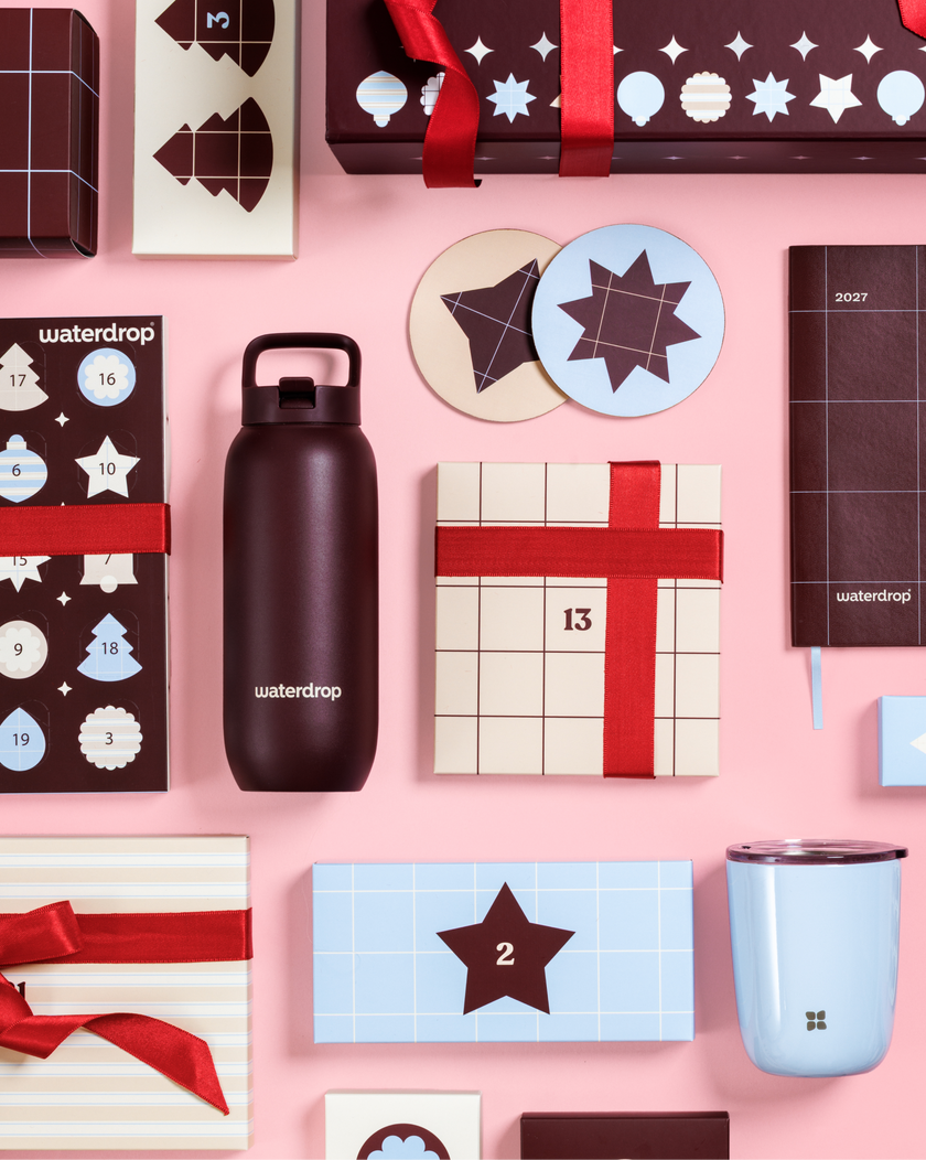 Calendari dell'Avvento e scatole regalo con nastro rosso su sfondo rosa, accanto a una bottiglia nera e a una tazza azzurra.
