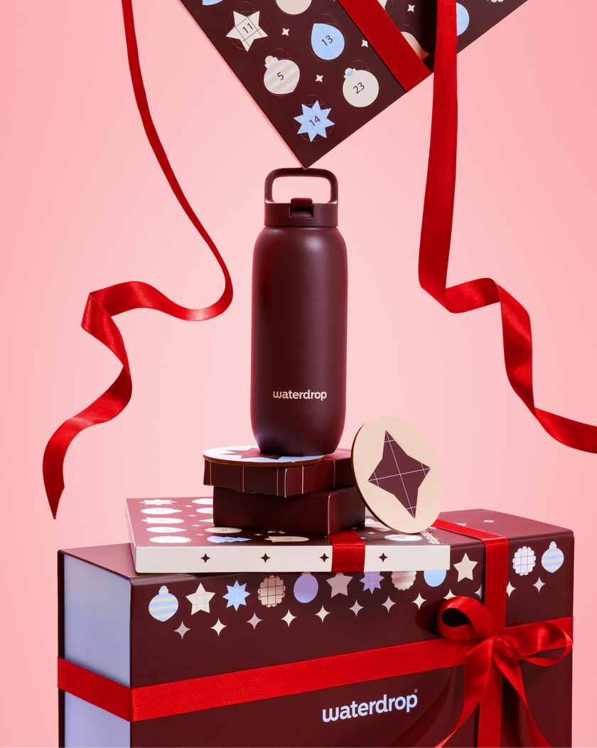 Bottiglia d'acqua nera con coperchio a anello su scatole regalo impilate legate con nastri rossi su sfondo rosa.