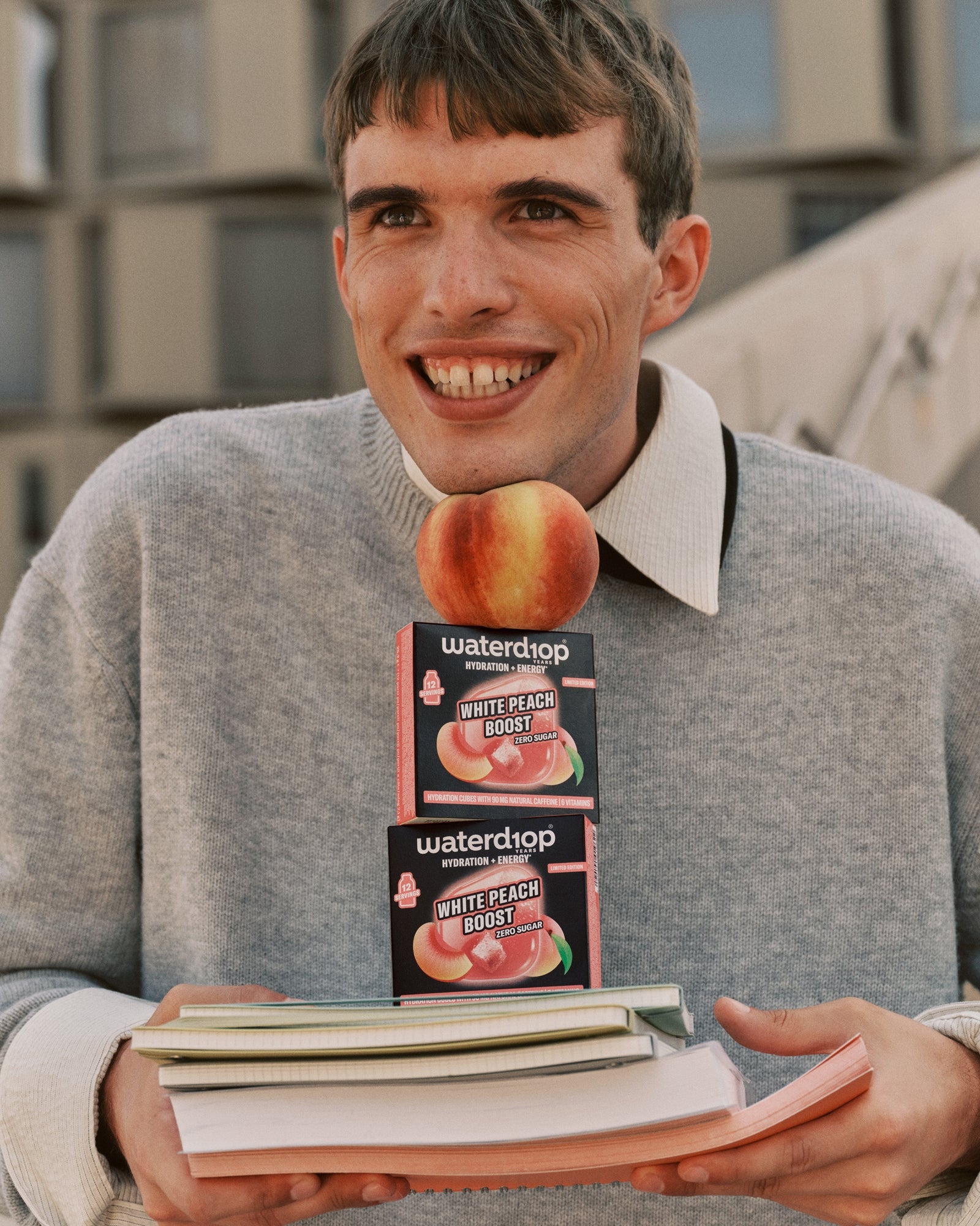 Giovane uomo in pullover grigio che tiene due confezioni al gusto pesca su una pila di libri.