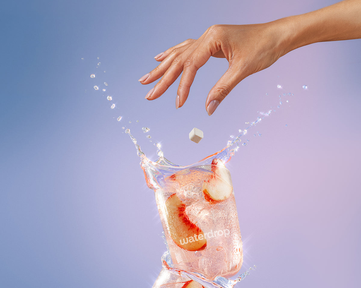 Una mano fa cadere un piccolo cubetto di ghiaccio in un bicchiere d'acqua con ghiaccio, causando schizzi.