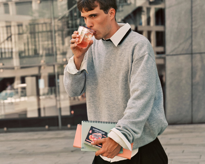 Uomo con maglione grigio che beve da un bicchiere mentre tiene un taccuino all’aperto in un contesto urbano.
