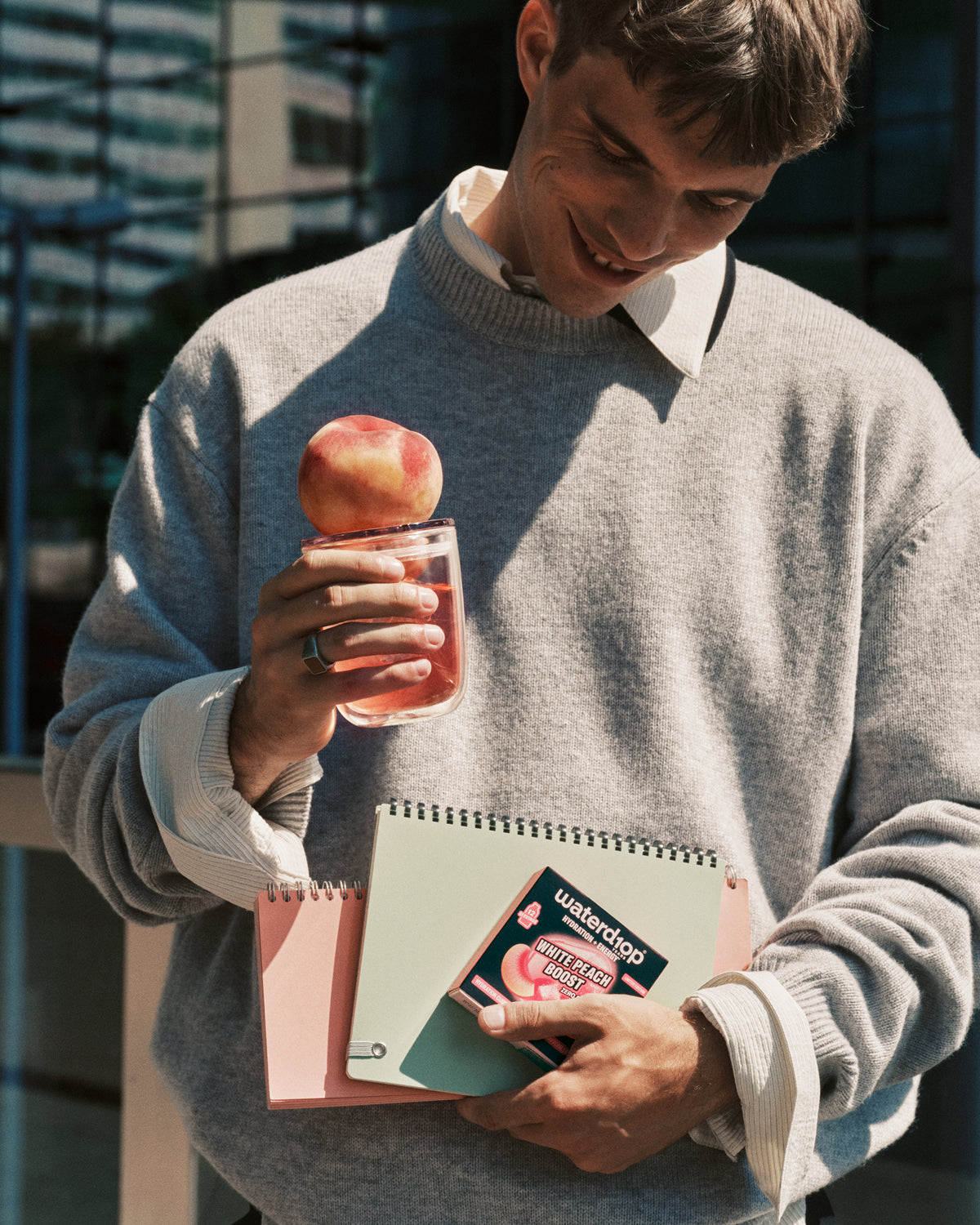 Uomo con maglione grigio all'aperto tiene una bevanda rosa con una mela sul bordo e un quaderno nella mano opposta.