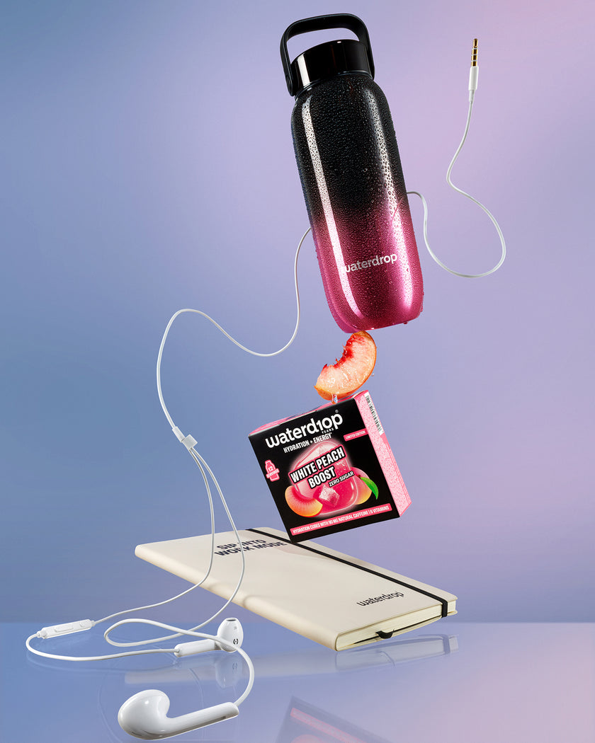Esposizione fluttuante di una bottiglia d'acqua con gradiente nero-rosa sopra una power bank bianca, collegata da cavi bianchi con una fetta di pesca nelle vicinanze.