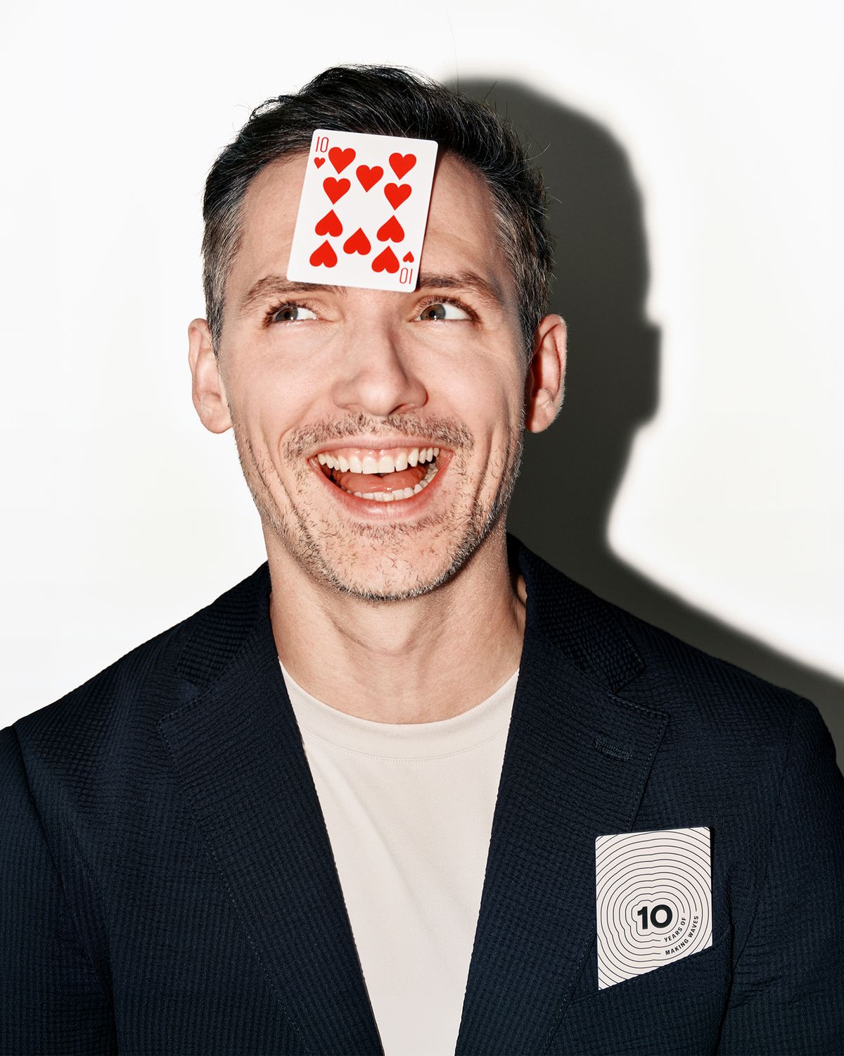 Uomo sorridente in blazer scuro e camicia chiara con una carta da gioco sulla fronte e un'altra in una tasca del petto.