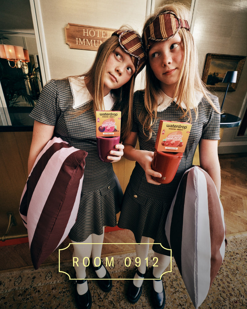 Due ragazze in abiti coordinati con cerchietti a quadri tengono drink colorati in un lobbby d'albergo, in piedi fianco a fianco con borse a righe.