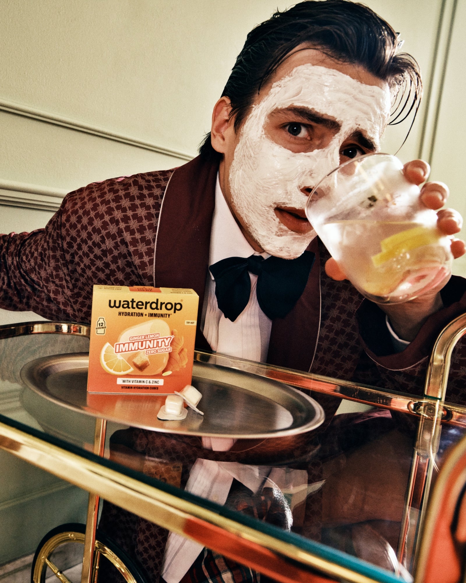Uomo in completo marrone con maschera per il viso che beve da un bicchiere al bancone.