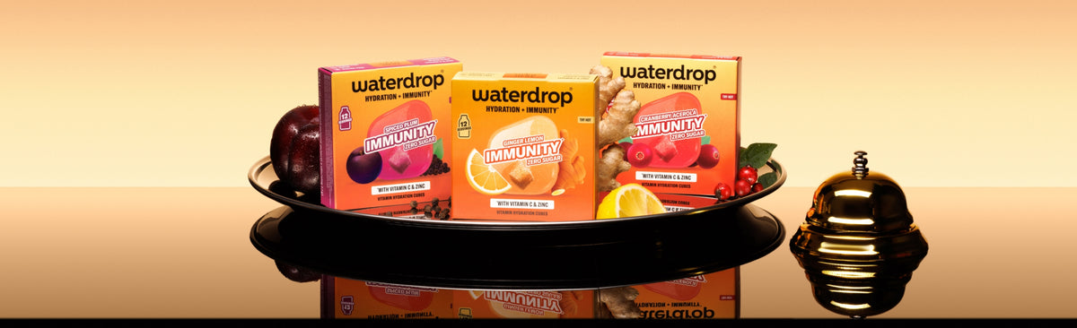 Vassoio con tre confezioni Waterdrop su una superficie riflettente e una piccola bottiglia decorativa a destra.
