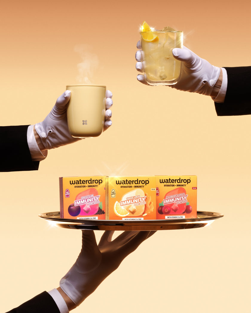 Vassoio trasportato da mani guantate presenta tre confezioni Waterdrop e due tazze, una delle quali con una fetta di limone.