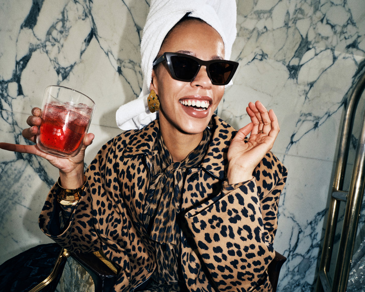 Donna con cappotto leopardato e occhiali da sole, asciugamano bianco in testa, tiene una bevanda rossa in un bicchiere davanti a una parete di marmo.