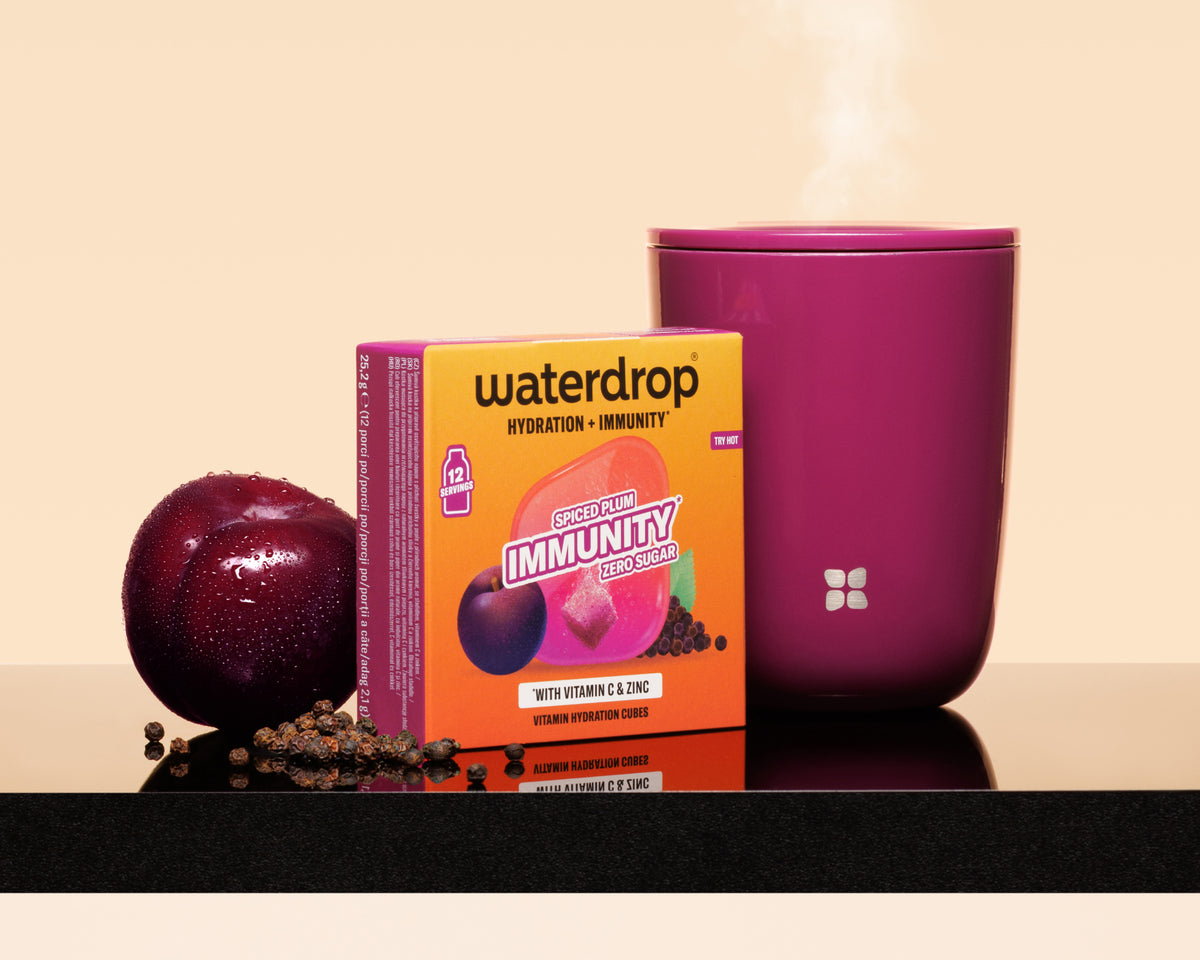 Tazza viola fumante accanto a una scatola arancione Waterdrop, una prugna e semi sparsi su una superficie riflettente.