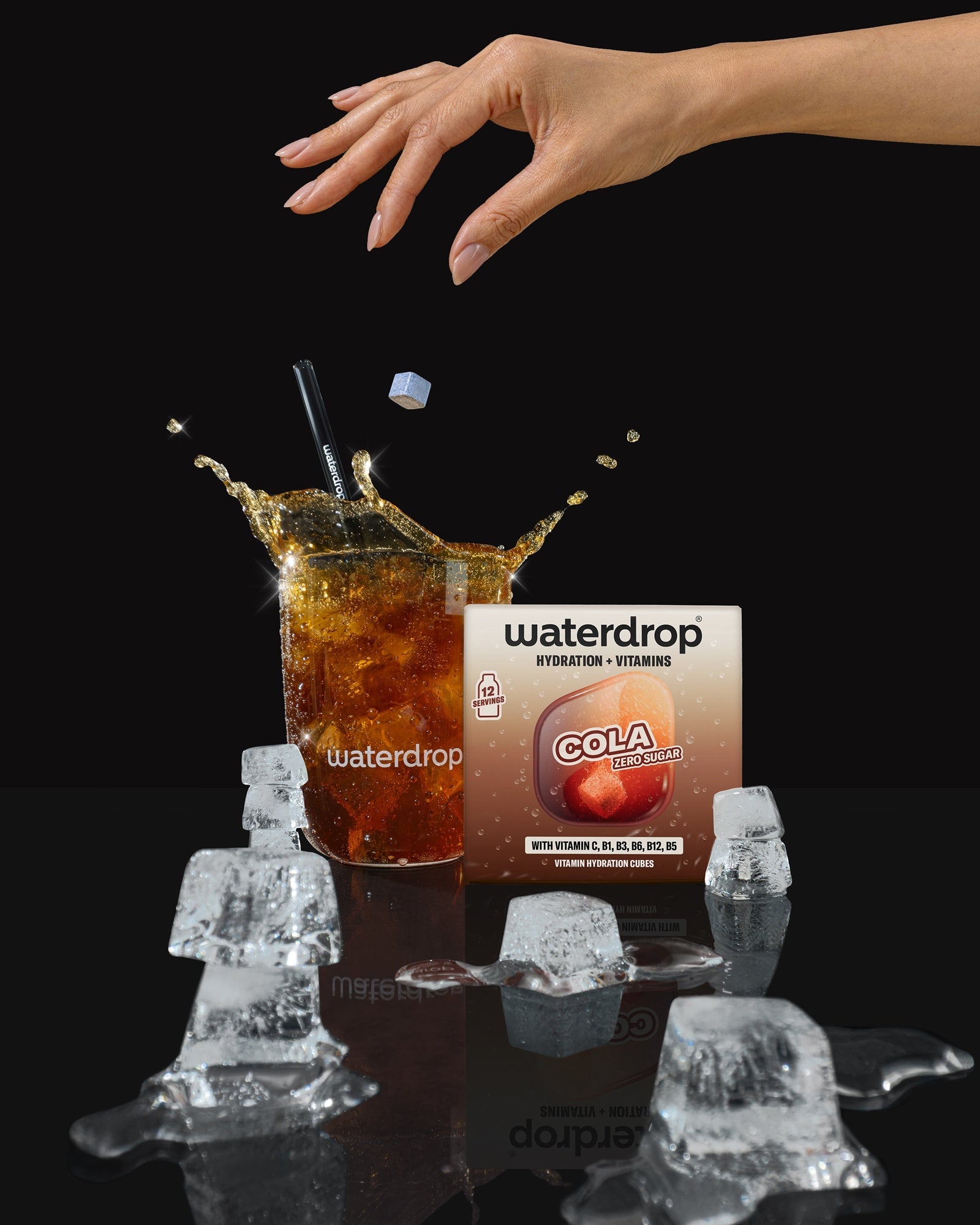 Una mano si avvicina a un bicchiere di cola con cubetti di ghiaccio, sullo sfondo una confezione di bevanda.