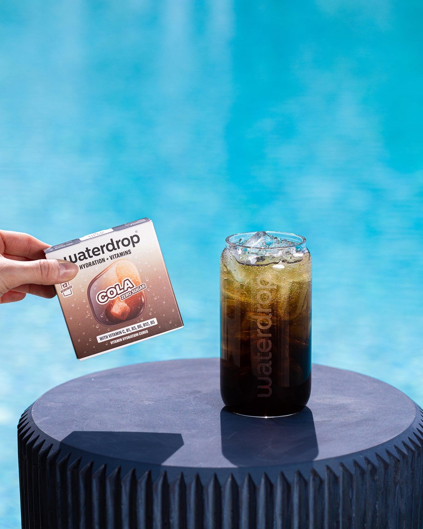 Una mano tiene una piccola confezione accanto a un bicchiere di cola ghiacciata su un supporto blu rotondo vicino alla piscina.